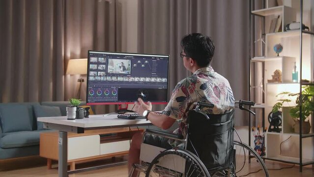 Back View Of Asian Man In Wheelchair Stretching While Editing And Color Grading The Video By A Desktop Next To The Camera At Home
