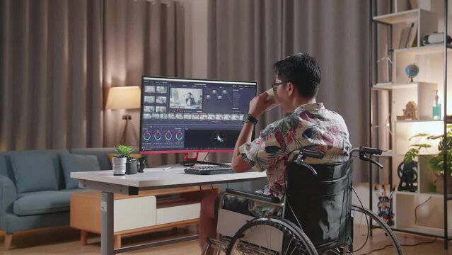 Back View Of Asian Man In Wheelchair Drinking Coffee While Editing And Color Grading The Video By A Desktop Next To The Camera At Home
