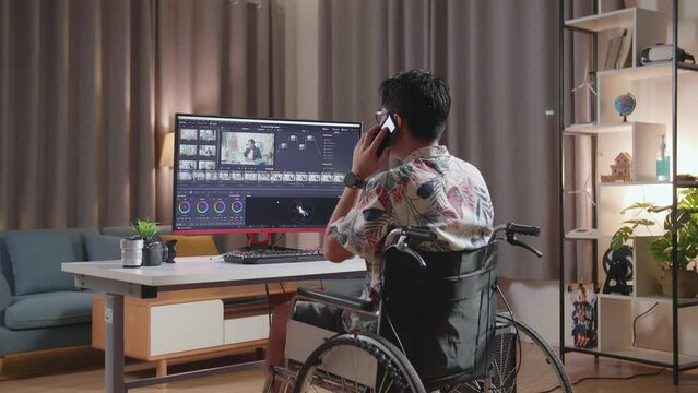 Back View Of Asian Man In Wheelchair Talking On Smartphone While Editing And Color Grading The Video By A Desktop Next To The Camera At Home
