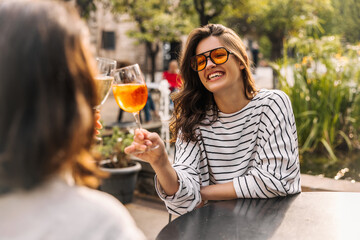 Joyful young caucasian girl with glass enjoys spending time in cafe summer. Brunette woman with wavy hair wears sunglasses. Relaxation concept