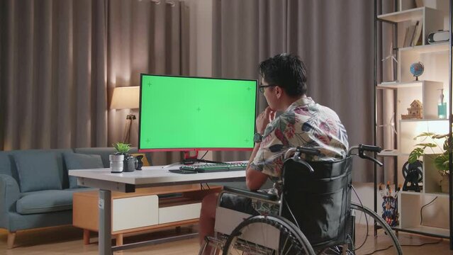 Back View Of Asian Man In Wheelchair Holding His Chin And Thinking About Something While Using Mock Up Green Screen Desktop Next To The Camera At Home
