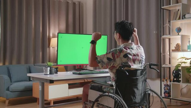 Back View Of Asian Man In Wheelchair Celebrating Succeed While Using Mock Up Green Screen Desktop Next To The Camera At Home
