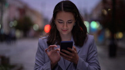 Portrait of a beautiful woman in a jacket using a smartphone on the street in the night city. Business lady stands with a phone on the background of blurred people and cars in the evening - 532854032
