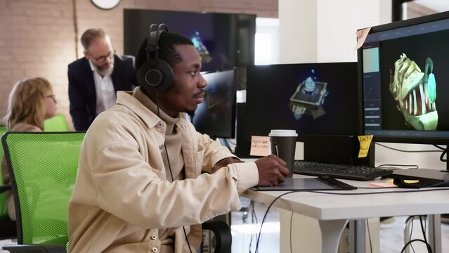 African American male artist designer draws on a tablet a 3D models objects for a computer game in the office of a project development studio.