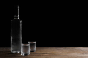 Bottle of vodka and shot glasses on wooden table against black background. Space for text