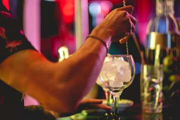 Closeup of hands of young bartender making a big cocktail with ice in a bar in the night with colorful lights