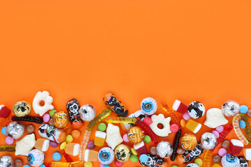 Orange background with copy space, halloween assorted candies at bottom - traditional eyeballs chocolates, jelly worms, ghosts. Happy Halloween holiday sale and trick-or-treat concept.