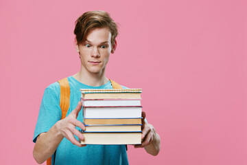 funny, red-haired student in a blue T-shirt and with an orange backpack on his back, holds a heavy stack of books in his hands and looks at the camera with surprise. Horizontal photo with empty space