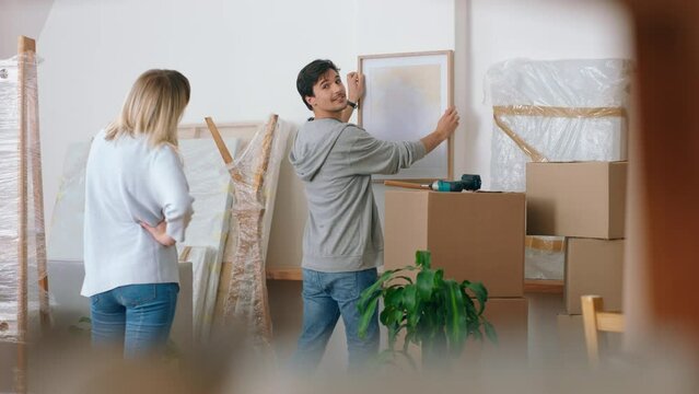 Home renovation, happy couple and interior decorating while moving in new house with cardboard boxes and hanging picture frame. Happy man and woman with mortgage approval helping decorate apartment
