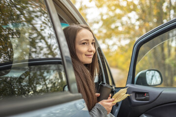 Woman is sitting in a car with a cup of coffee and bouquet of yellow autumn leaves, admiring the autumn landscape and enjoying the silence and beauty of nature