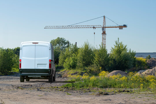 A white blank van parked on a new construction site with trees, bushes and bosk still growing, a crane erected in the background and copy space in the clear summer sky