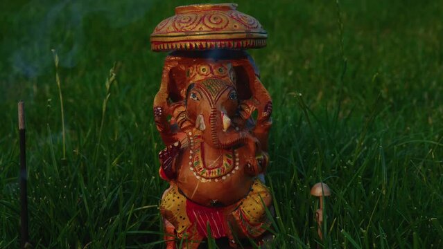 Close-up, a move backwards, Deity Sri Ganesha from sandalwood stands on green grass with a row of steaming incense and a growing mushroom.
