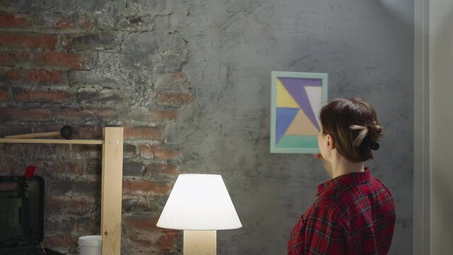 Woman seeks place for watercolor painting on grange style wall in restored living room. Interior designer works upon apartment decor. Home ornate trends