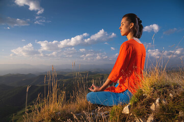 young caucasian woman meditates in the lotus position in the grass in the mountains near the cliff...