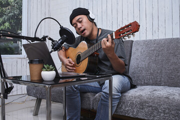 Asian guy playing the guitar recording audio podcast in studio using microphone and laptop. People...