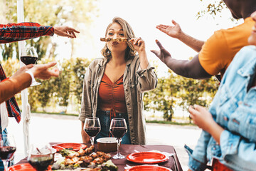 Friends having fun party pointing a curvy young woman making faces holding skewers grilled on...
