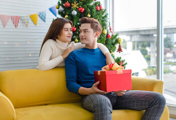 Obraz na płótnie Canvas Asian boyfriend sitting on cozy sofa holding opening red wrapped present gift box showing to happy girlfriend in home living room full decorated with pine trees celebrating Christmas eve festival