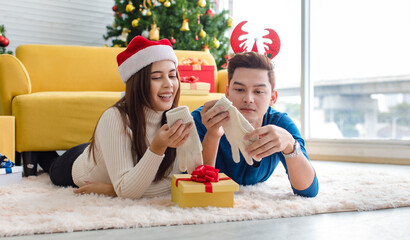 Asian cheerful boyfriend wears deer antler headband and girlfriend in sweater and red Santa Claus hat laying down on floor looking each other eyes in decorated living room celebrating Christmas eve