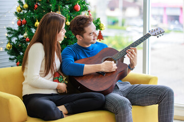 Asian happy cheerful romantic lover couple girlfriend smiling hugging cuddling boyfriend sitting on cozy sofa playing guitar sing song celebrating Christmas eve in full decorated home living room