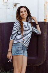 a girl with a dreadlocked hairstyle poses in the summer outdoors, dressed in a T-shirt and denim shorts