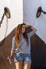 a girl with a dreadlocked hairstyle poses on the beach near a ship in summer, a sunny day, dressed in a T-shirt and denim shorts