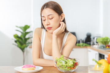 Obraz na płótnie Canvas Diet, Dieting asian young woman confused choose, choice eating green salad in bowl, looking at pink sweet doughnut at home, eat food good health when hungry. Weight loss person, temptation concept.