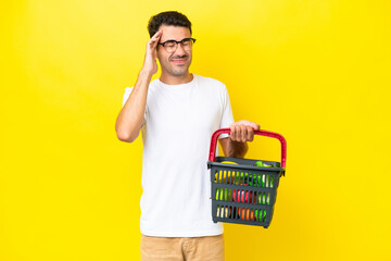 Young handsome man holding a shopping basket full of food over isolated yellow background with...