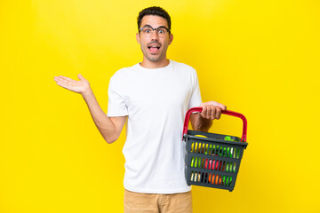 Young handsome man holding a shopping basket full of food over isolated yellow background with...