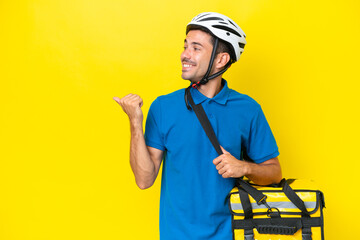 Young handsome man with thermal backpack over isolated yellow background pointing to the side to present a product