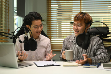 Two smiling asian man podcasters using laptop and condenser microphone to recording podcast in home studio