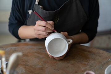 Fototapeta na wymiar Craftswoman creates new masterpiece in work studio making sketch of future pattern on white mug. Lady master wants to decorate kitchen with painted clay dish using pencil to draw interesting pattern