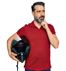 Middle aged man with beard holding motorcycle helmet serious face thinking about question with hand...