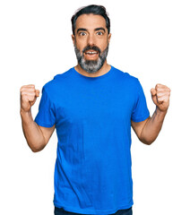 Middle aged man with beard wearing casual blue t shirt celebrating surprised and amazed for success with arms raised and open eyes. winner concept.
