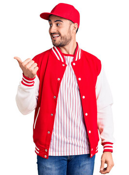 Young handsome man wearing baseball jacket and cap smiling with happy face looking and pointing to the side with thumb up.