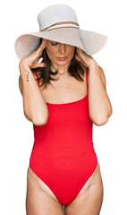 Beautiful brunette woman wearing swimsuit and summer hat with hand on head for pain in head because stress. suffering migraine.