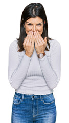 Beautiful brunette woman wearing casual clothes laughing and embarrassed giggle covering mouth with...