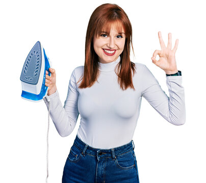 Redhead young woman holding electric steam iron doing ok sign with fingers, smiling friendly gesturing excellent symbol