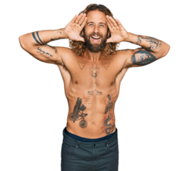 Handsome man with beard and long hair standing shirtless showing tattoos smiling cheerful playing peek a boo with hands showing face. surprised and exited