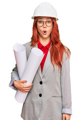 Young redhead architect woman wearing safety helmet holding blueprints scared and amazed with open mouth for surprise, disbelief face