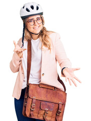 Young caucasian woman wearing bike helmet and leather bag clueless and confused expression with arms and hands raised. doubt concept.
