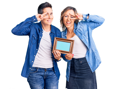 Couple of women holding empty frame doing peace symbol with fingers over face, smiling cheerful showing victory