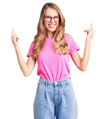 Young beautiful caucasian woman with blond hair wearing casual clothes and glasses shouting with crazy expression doing rock symbol with hands up. music star. heavy concept.