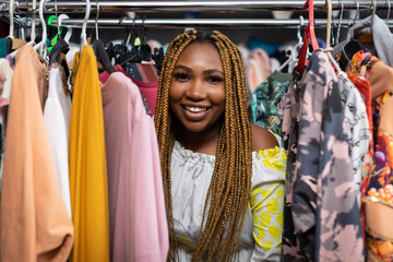 A smiling dark-skinned woman leans out from behind a rack full of clothing.