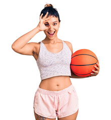 Young woman holding basketball ball smiling happy doing ok sign with hand on eye looking through fingers