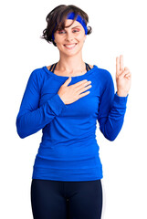 Beautiful young woman with short hair wearing training workout clothes smiling swearing with hand...