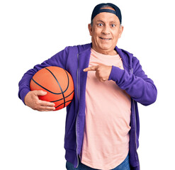 Senior handsome grey-haired man holding basketball ball smiling happy pointing with hand and finger
