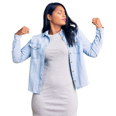 Hispanic woman with long hair wearing casual denim jacket showing arms muscles smiling proud. fitness concept.