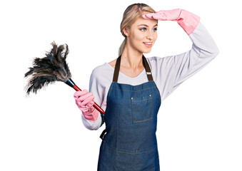 Beautiful young blonde woman wearing apron holding cleaning duster very happy and smiling looking far away with hand over head. searching concept. - Powered by Adobe
