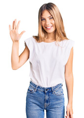 Beautiful caucasian woman with blonde hair wearing casual white tshirt showing and pointing up with fingers number five while smiling confident and happy.