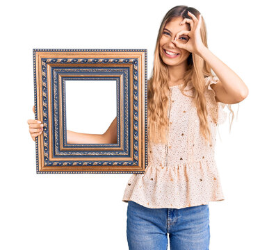 Beautiful caucasian woman with blonde hair holding empty frame smiling happy doing ok sign with hand on eye looking through fingers